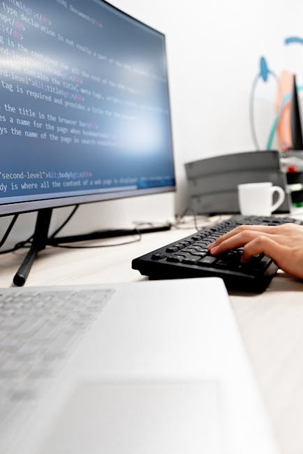 A person using a computer for gardening-related research or planning, with the keyboard in their hand and a monitor displaying lines of code or digital content. The workspace features a white desk wit