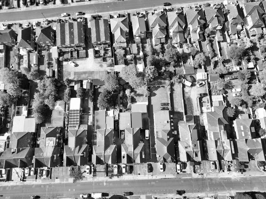 An aerial view of a residential suburban garden area, showing a row of closely spaced houses with pitched roofs and small backyards. The gardens feature a variety of plants, trees, and well-maintained