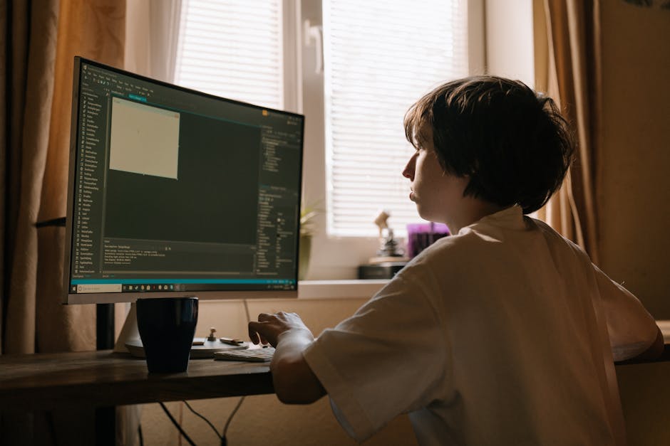 A woman with short dark hair and light skin sits at a wooden desk in a room illuminated by natural light from a window behind her. She is focused on a large, widescreen computer monitor displaying a d